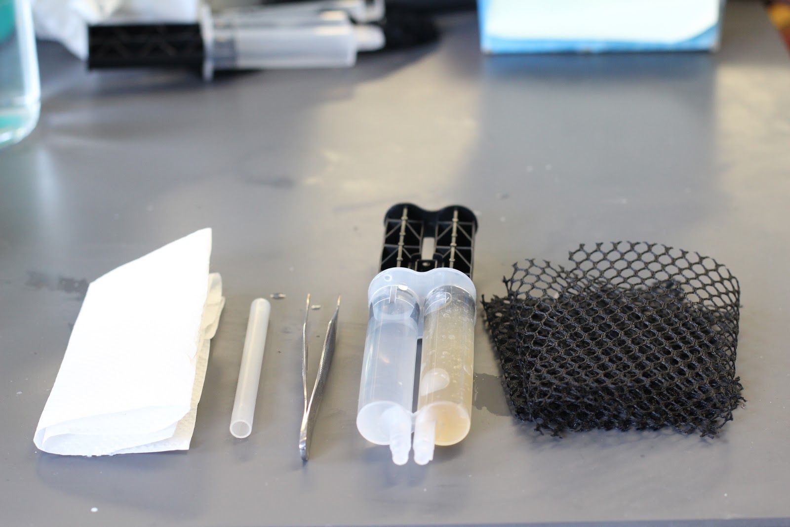 A photo of the GelAid components, including a tube, dual tubes with liquid in one and gel in the other, tweezers, and a black netting